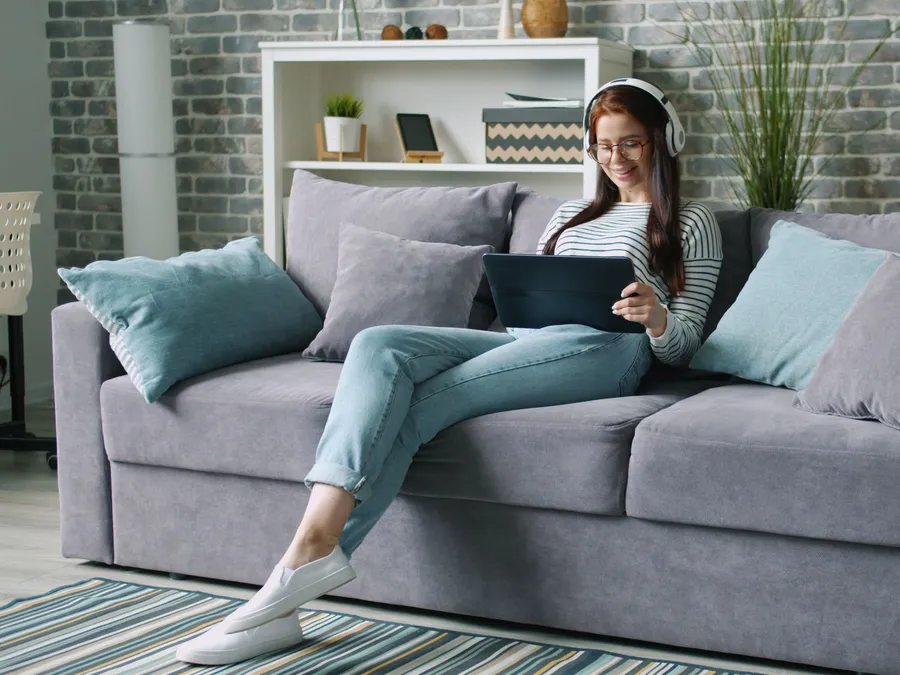 Woman wearing headphones and glasses relaxing on a gray sofa while watching a video on a tablet