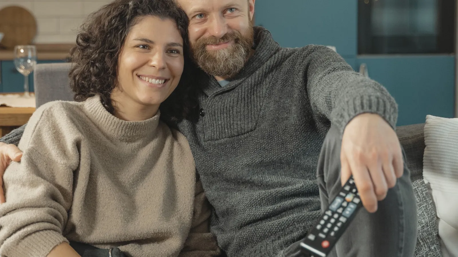 Smiling couple in sweaters relaxing together on a sofa at home while watching TV, one of them holding a remote control